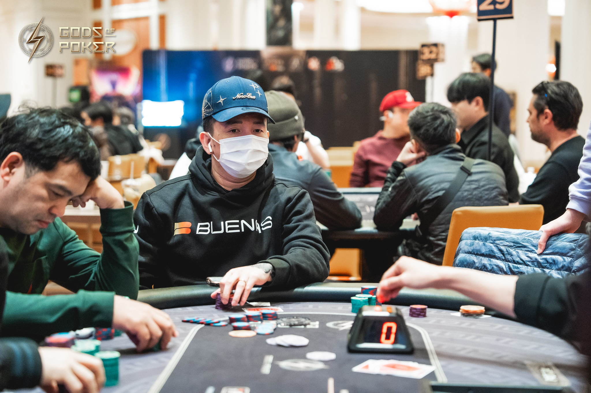 A man wearing a mask and cap playing poker at a busy casino table.