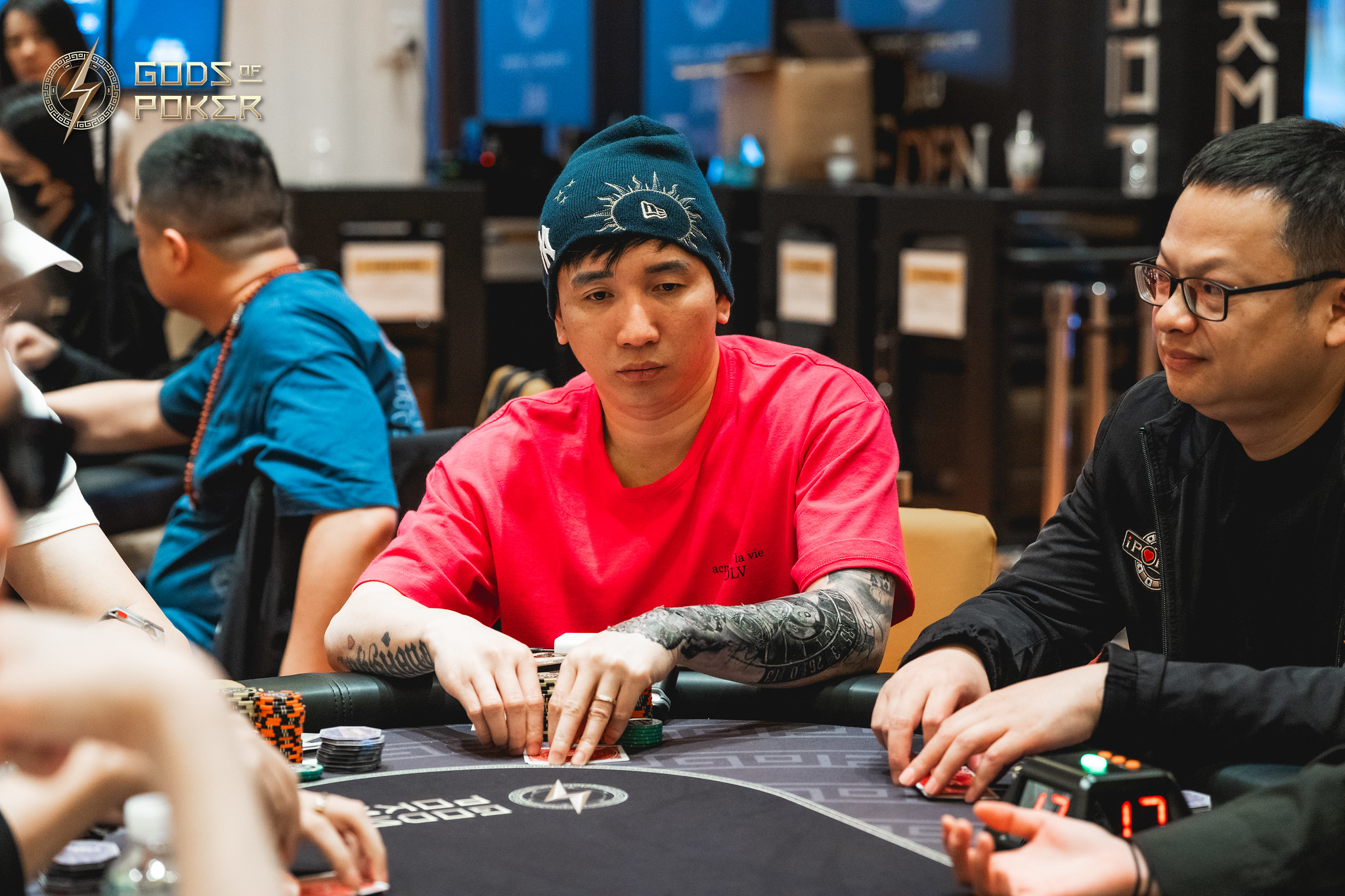 A man in a red shirt and beanie with tattooed arms focused on playing poker.