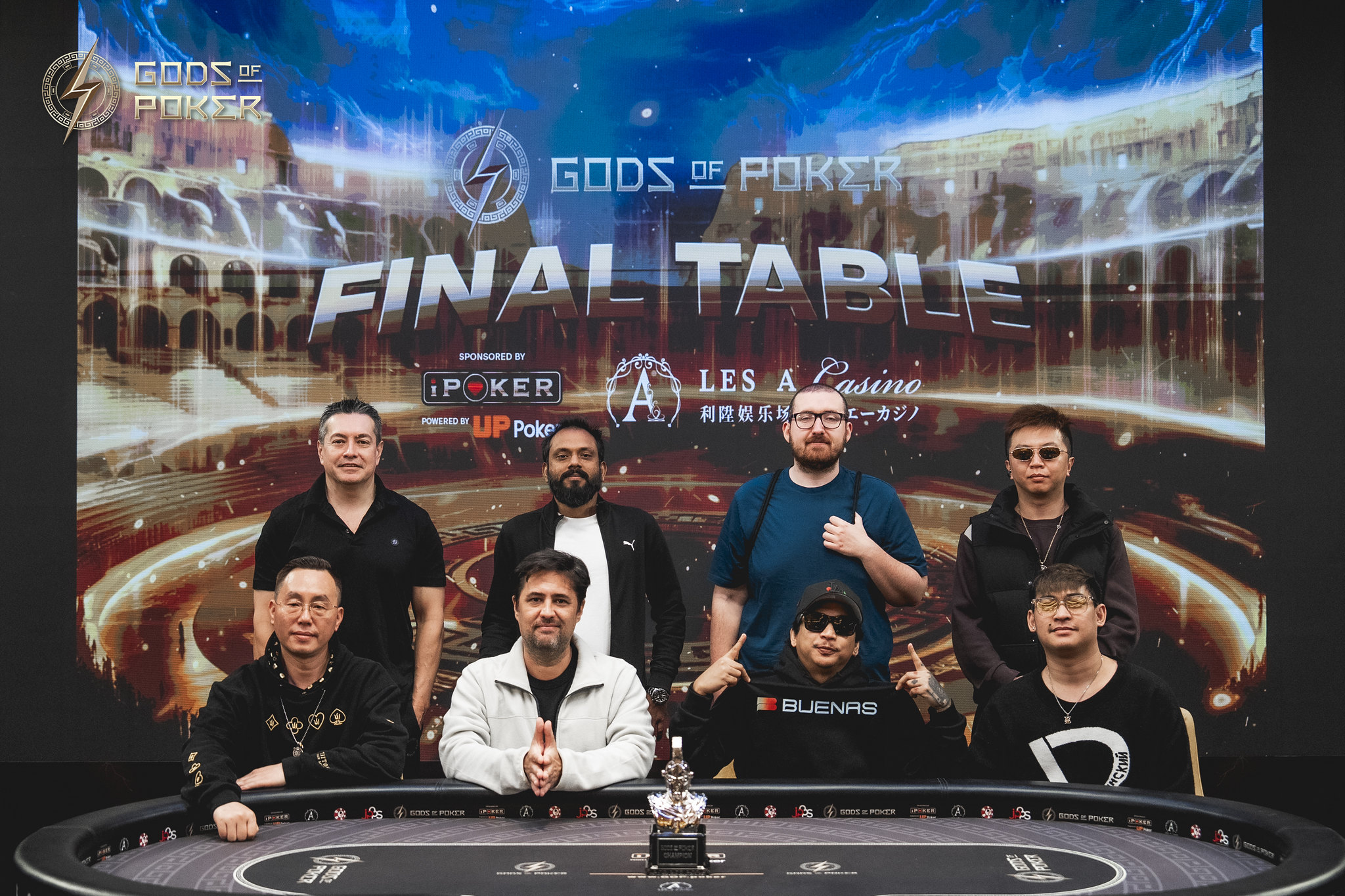 Eight male poker players posing at a final table with a trophy.