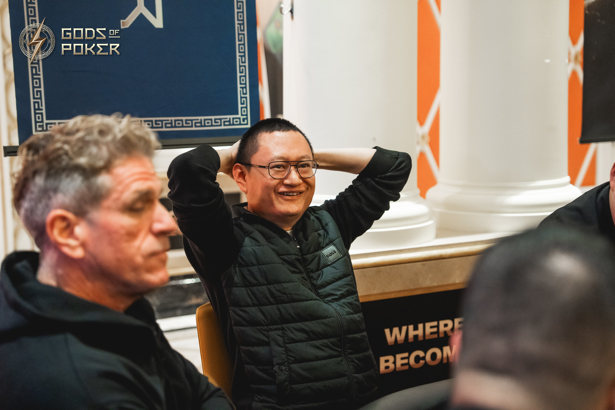 A smiling man in glasses leans back with hands behind his head at an event.