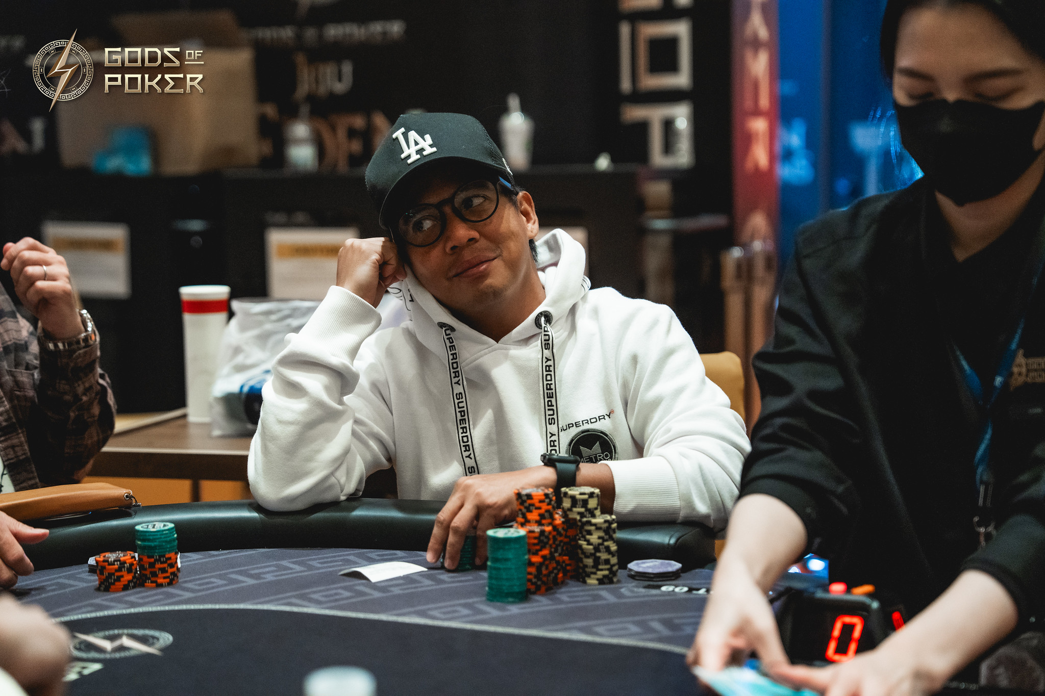 Man in a white hoodie and cap smiles while playing poker at a table.