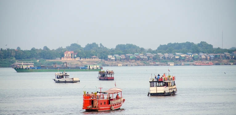 Diverse boats, including passenger and cargo vessels, navigate a wide river near a distant shore.