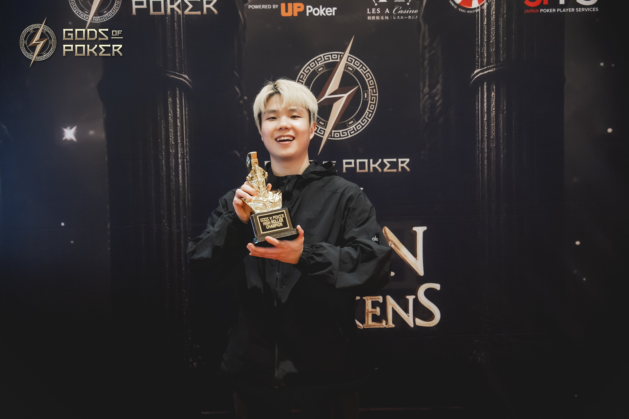 A smiling young person with light hair proudly holds a golden trophy at a poker event.