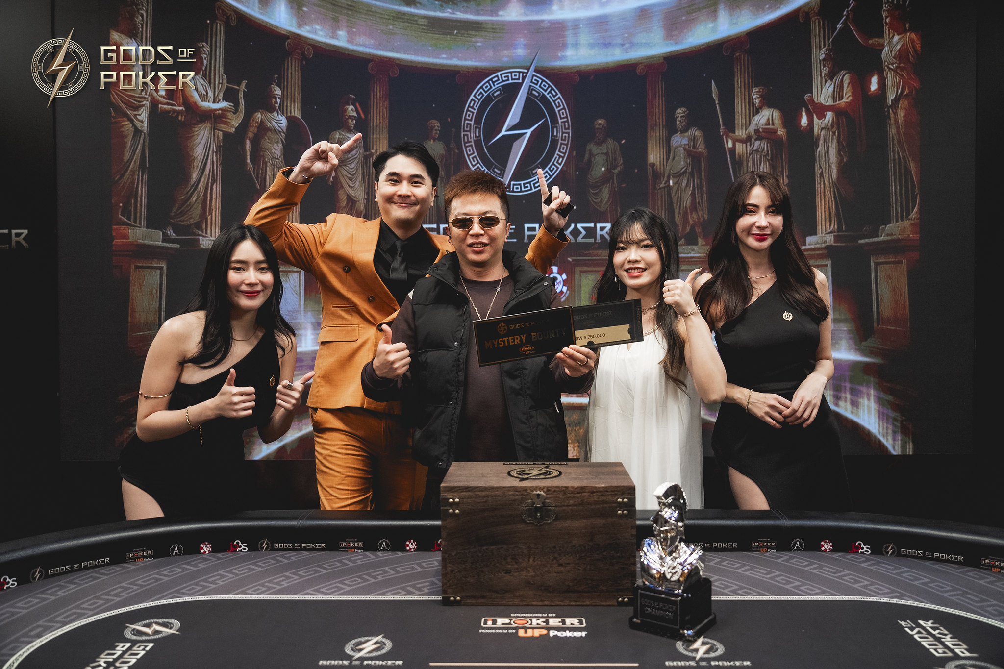 Five people celebrating a poker tournament victory with trophies and a large screen.