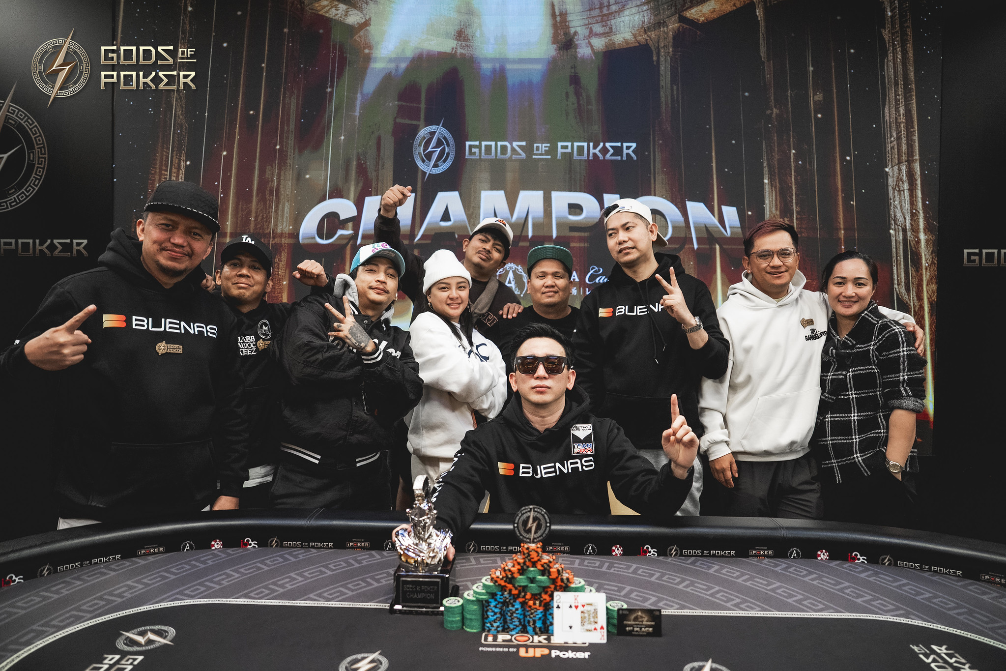 A poker champion sits at a table with a trophy, surrounded by a cheering team.
