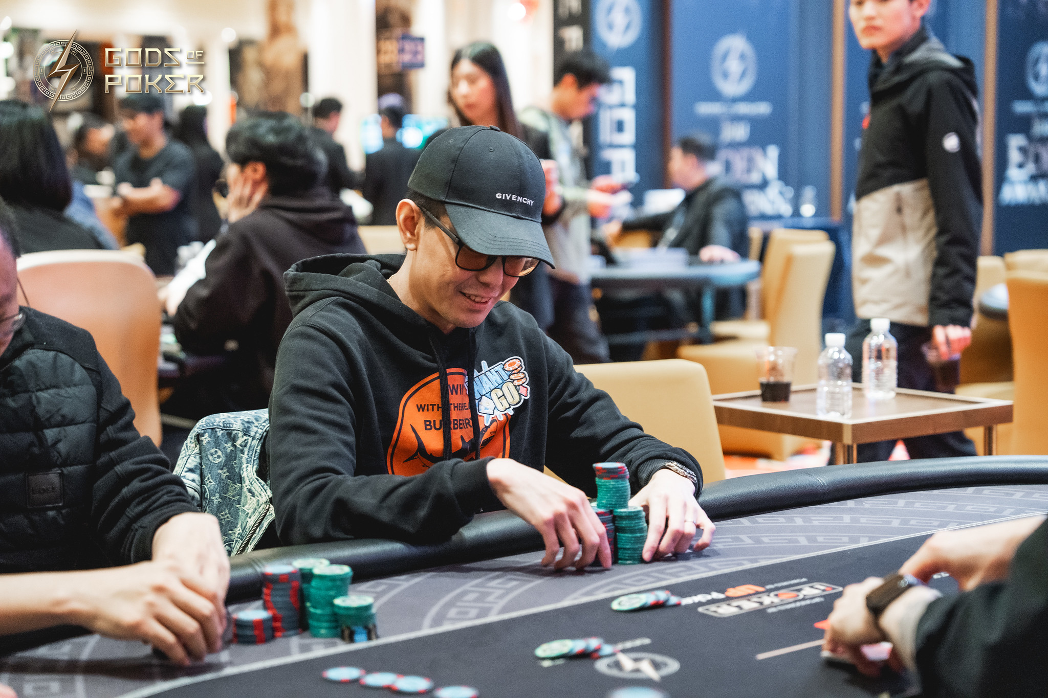 A man wearing a black cap and hoodie stacks green poker chips at a table during a game.
