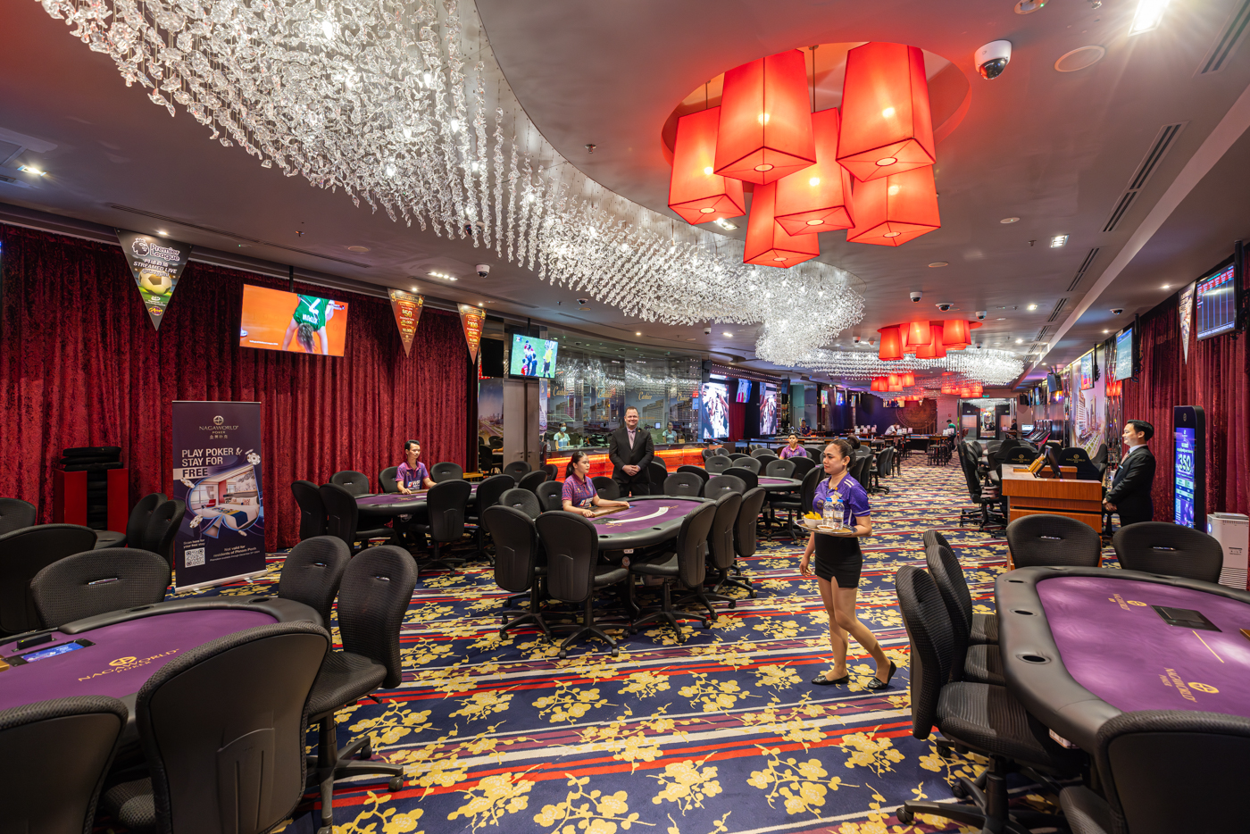 Luxurious casino interior with multiple poker tables, ornate chandeliers, and a server walking.