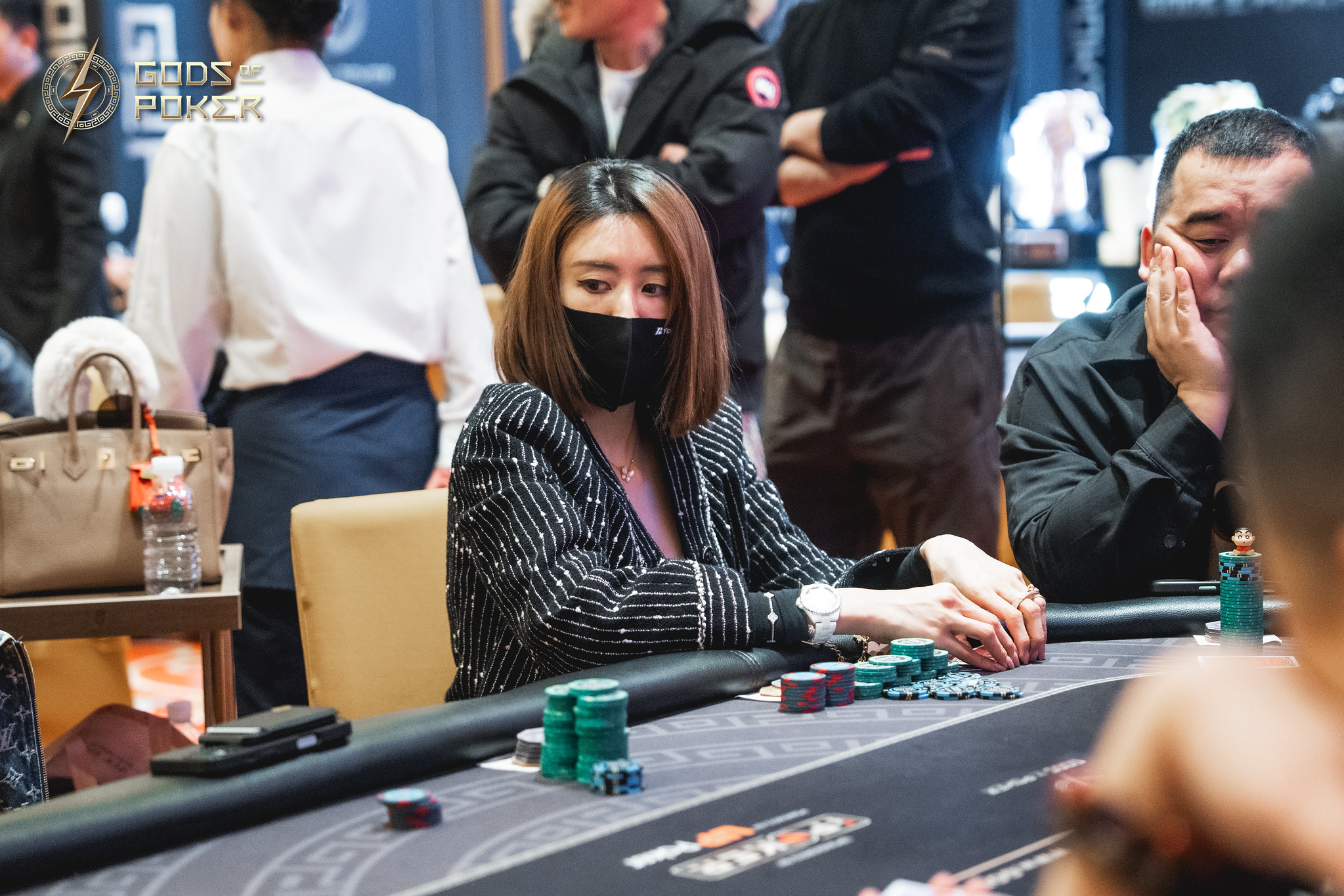 A woman in a black mask intently plays poker at a table with chips.