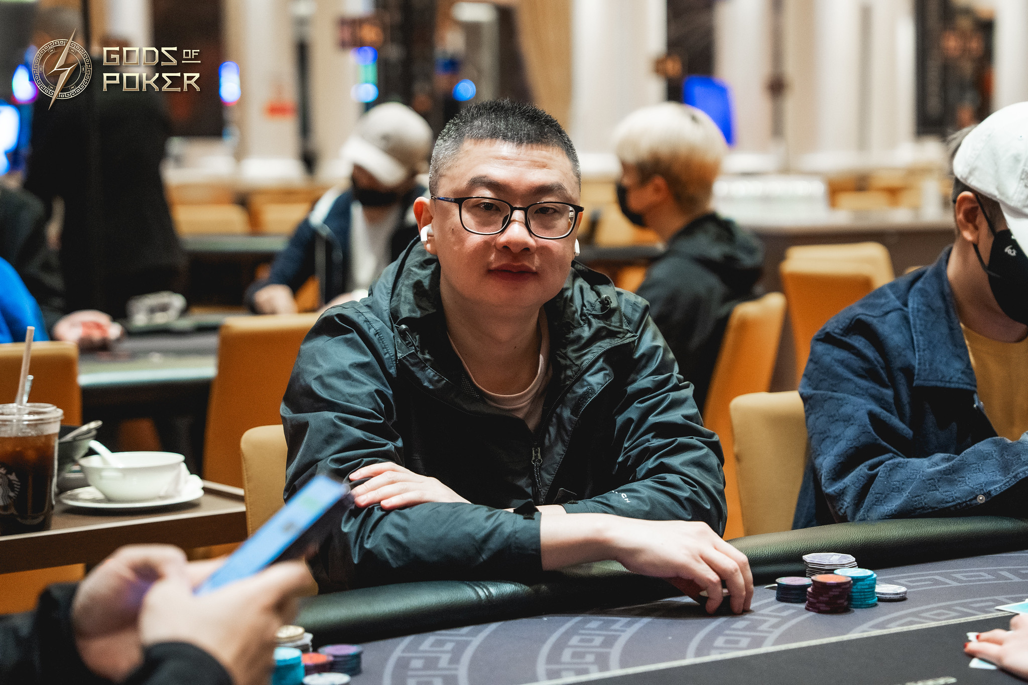 Man in glasses and dark jacket smiles at a poker table with chips.