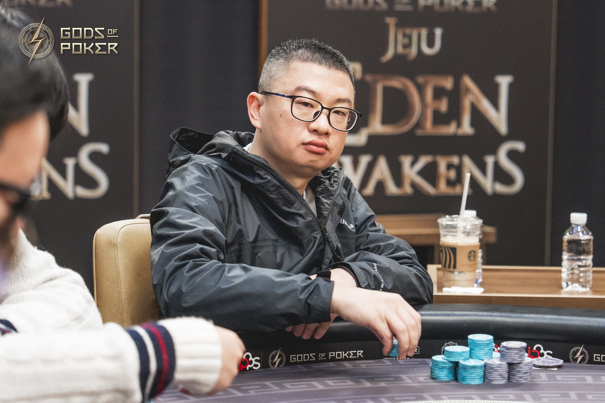 A serious man in glasses sits at a poker table with a stack of blue chips.