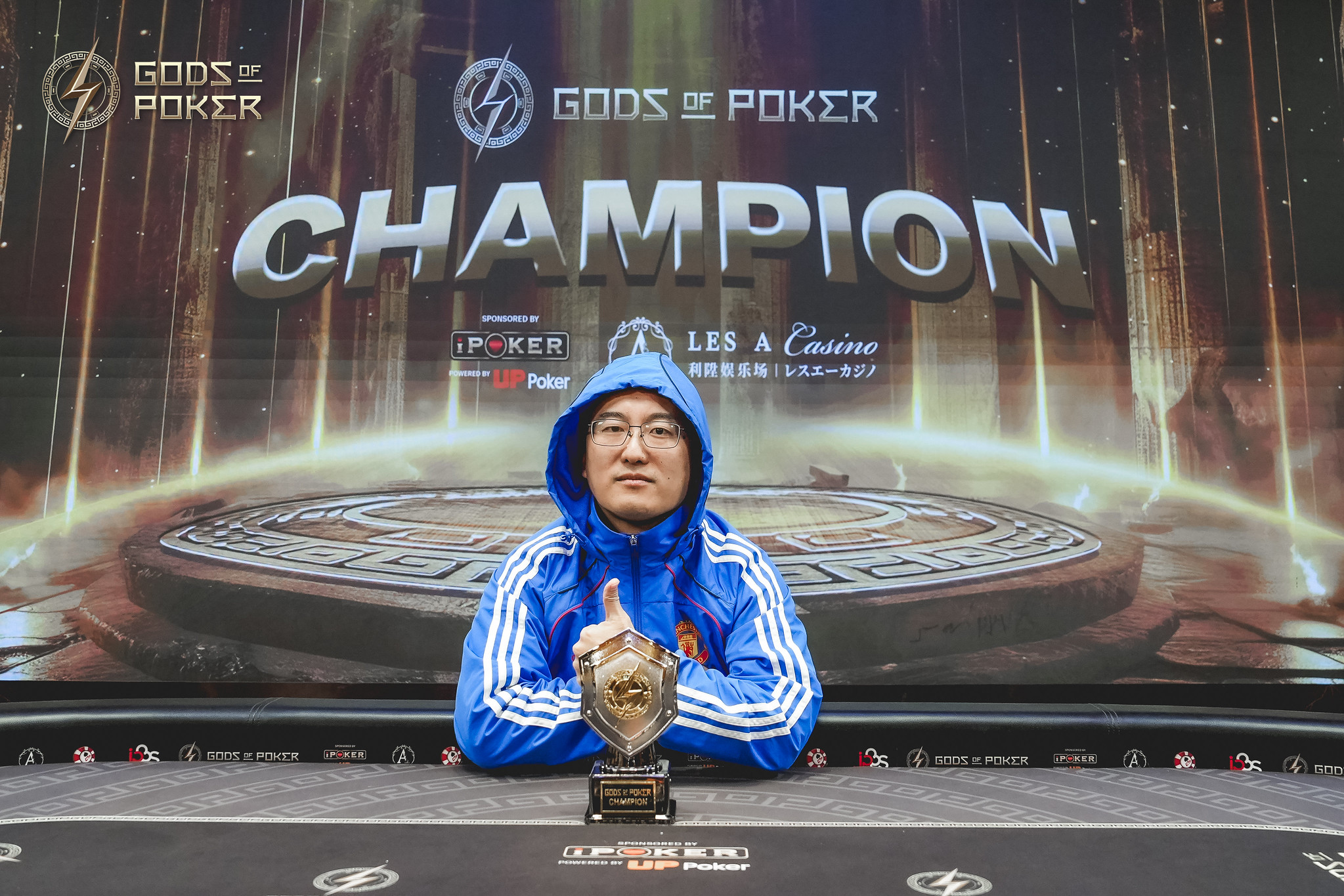 Man in blue jacket holds a poker trophy and gives a thumbs-up after winning.