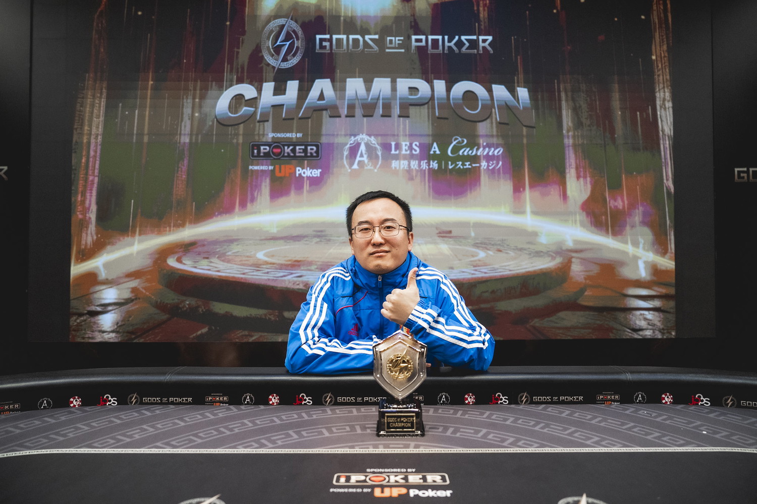 A male poker champion in a blue tracksuit giving a thumbs-up behind his trophy.