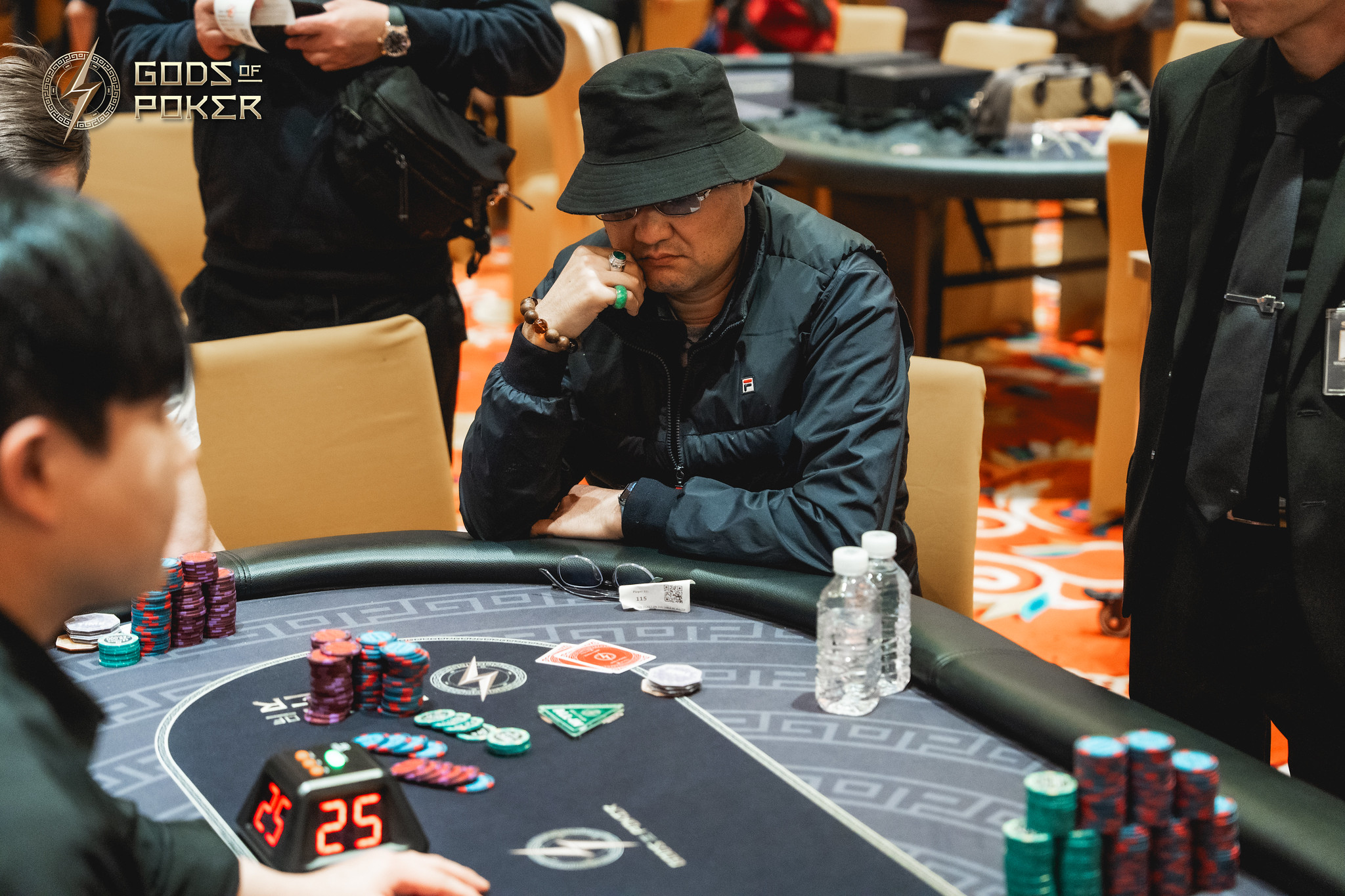 A man in a black bucket hat sits intently at a poker table, contemplating his cards.