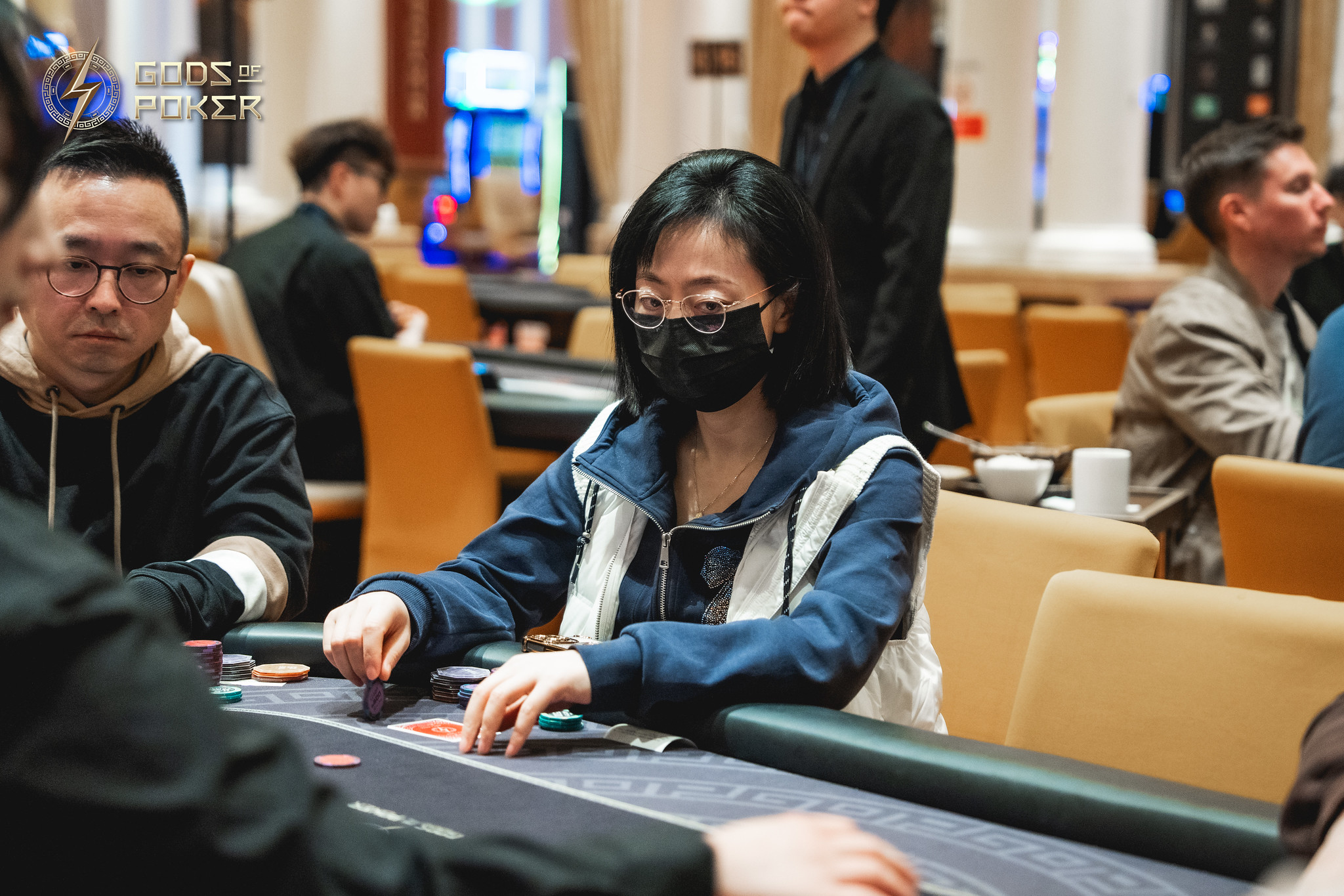 A woman in a face mask and hoodie plays poker at a casino table.