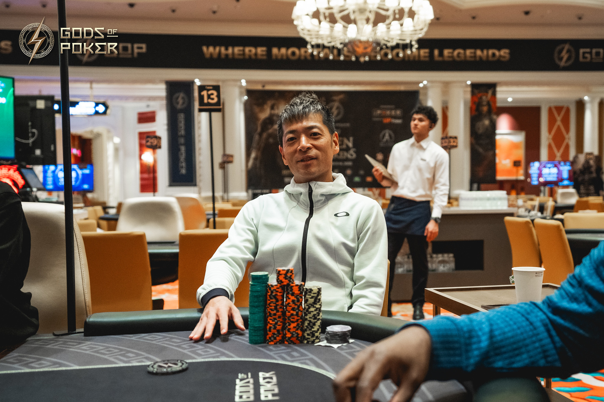 A man in a light jacket sits at a poker table with a stack of chips.
