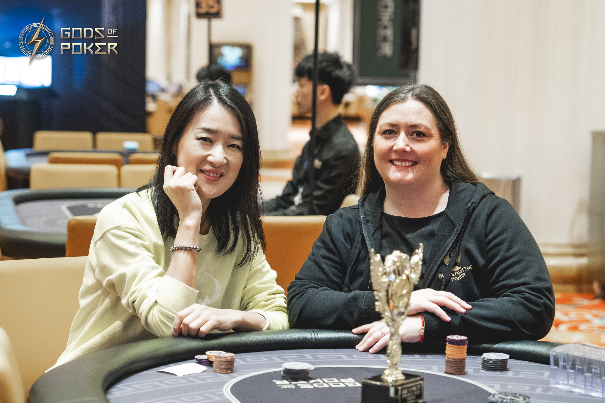 Two smiling women at a poker table, one proudly holding a golden trophy.