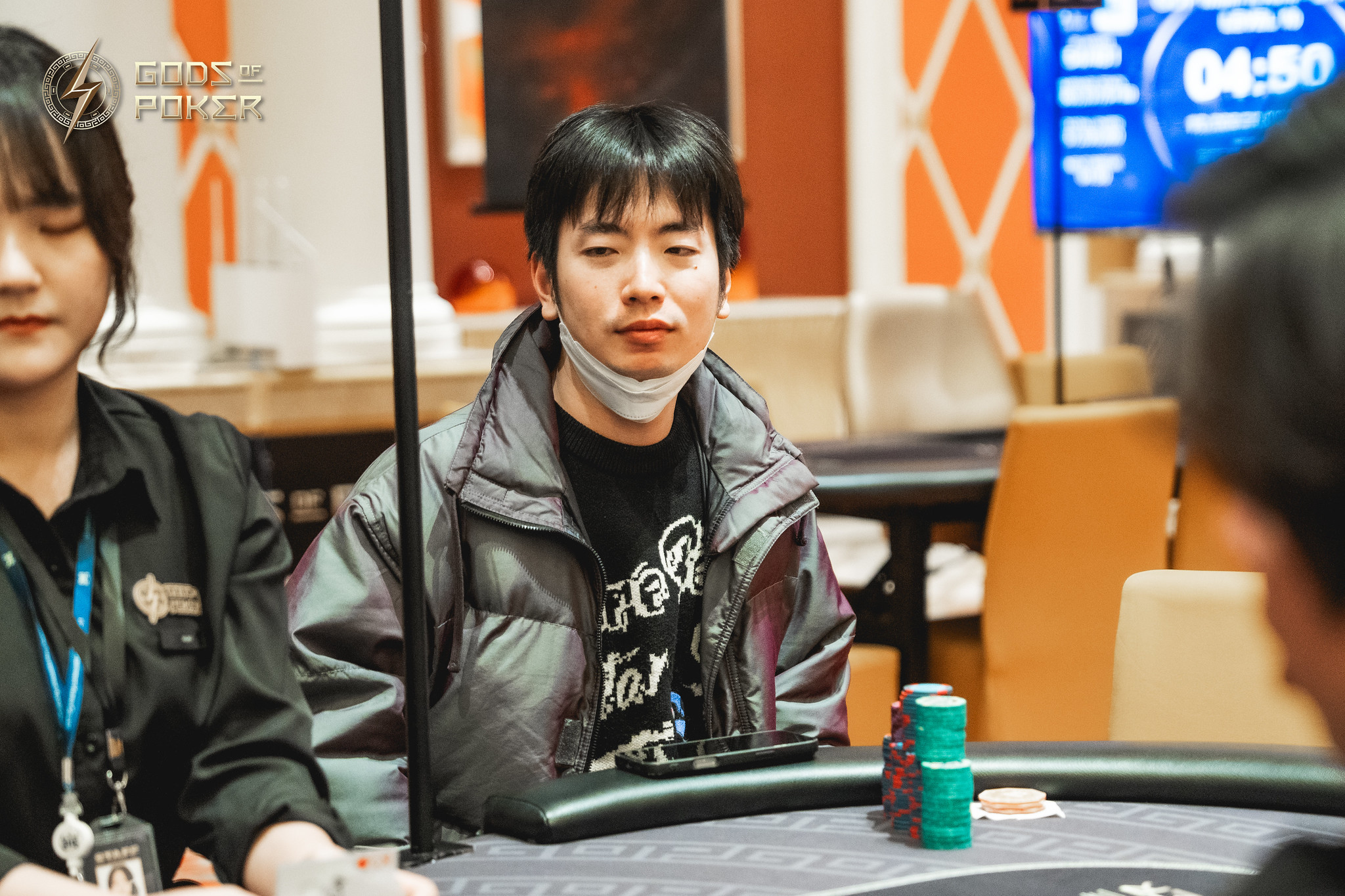 Young man in a grey puffer jacket sits pensive at a poker table.
