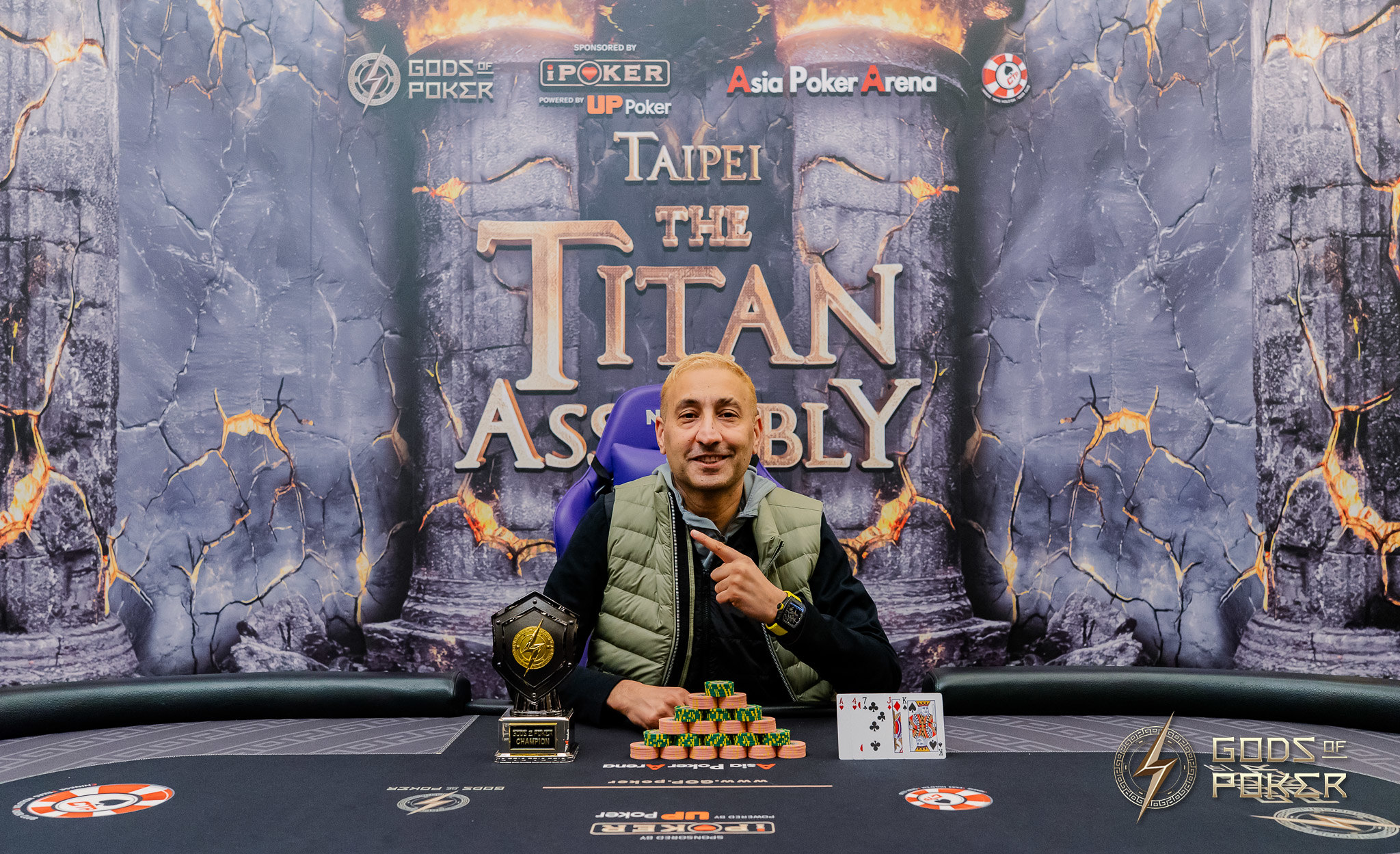 Smiling man points to poker chips and trophy at 'Taipei The Titan Assembly' event.