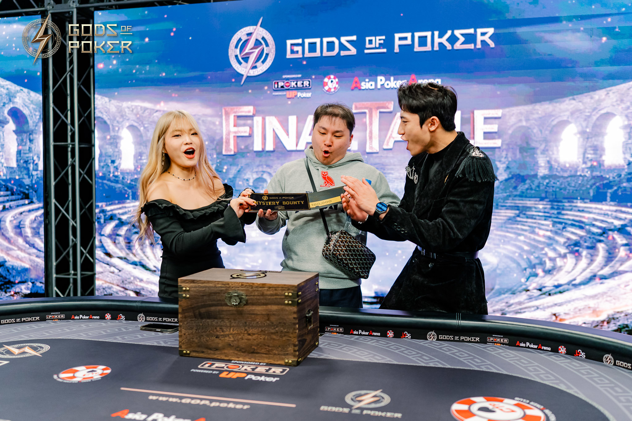 Three people at a poker final table, one man holding a key over a mysterious wooden treasure chest.