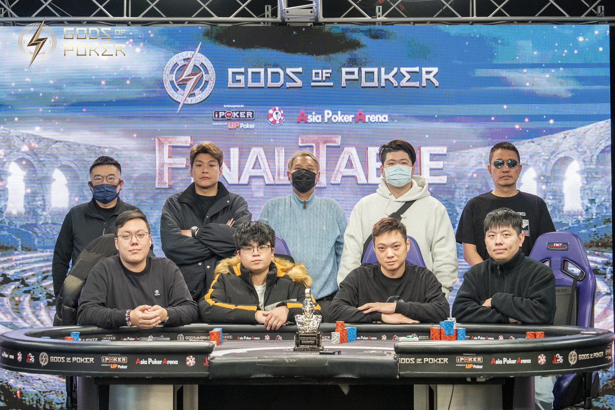 Eight poker players pose around a final table with 'Gods of Poker' branding in the background.