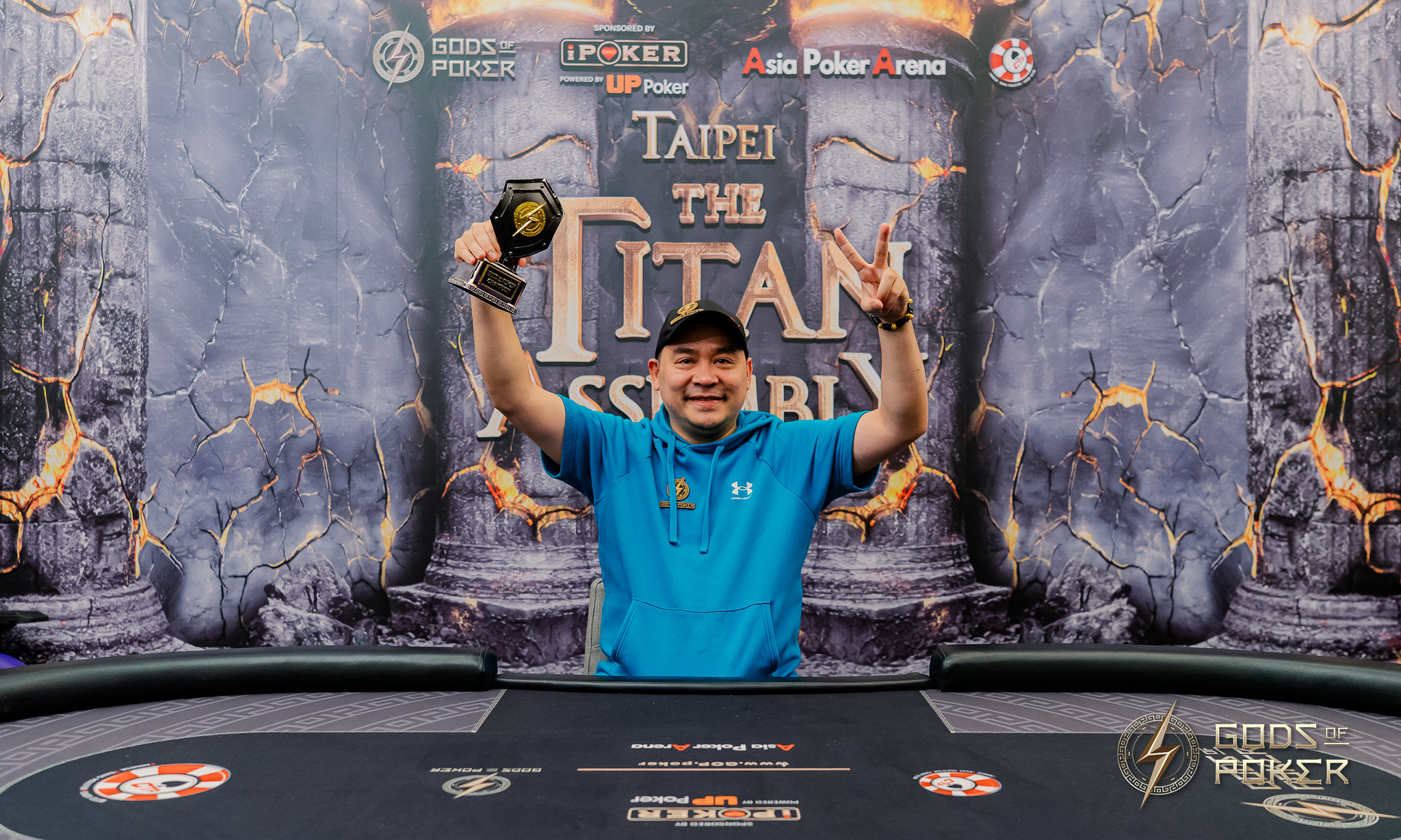 Smiling man celebrating poker victory with trophy and peace sign at Taipei Titan Assembly.