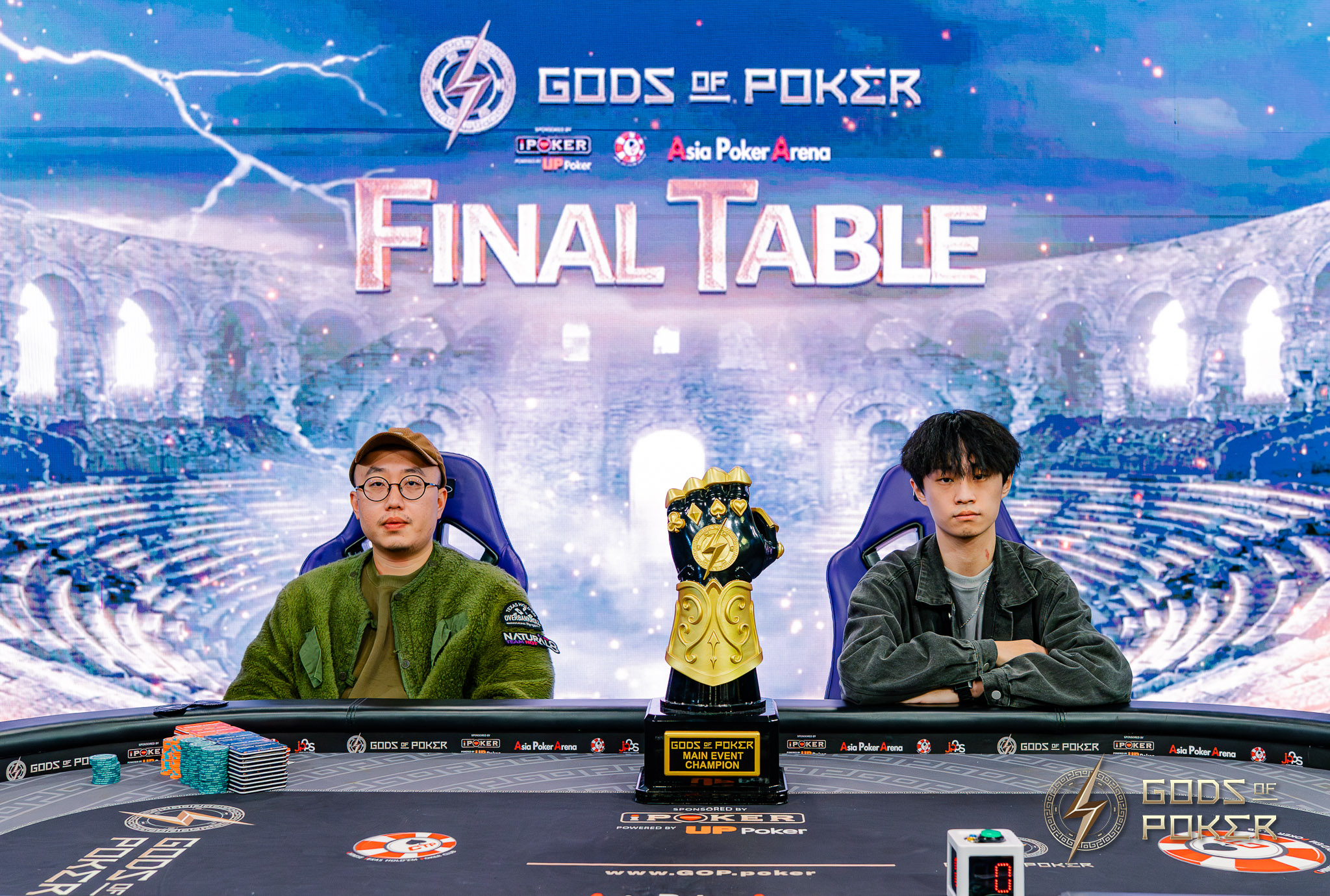 Two poker players sit at a final table with a large golden trophy between them.