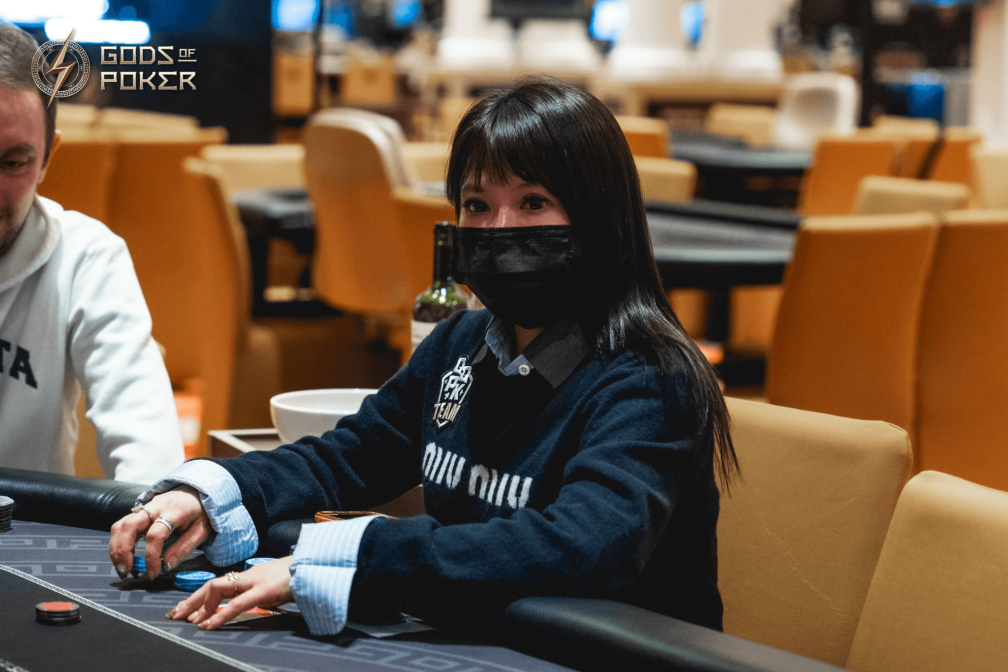 A woman in a black mask and dark sweater plays poker at a casino table.