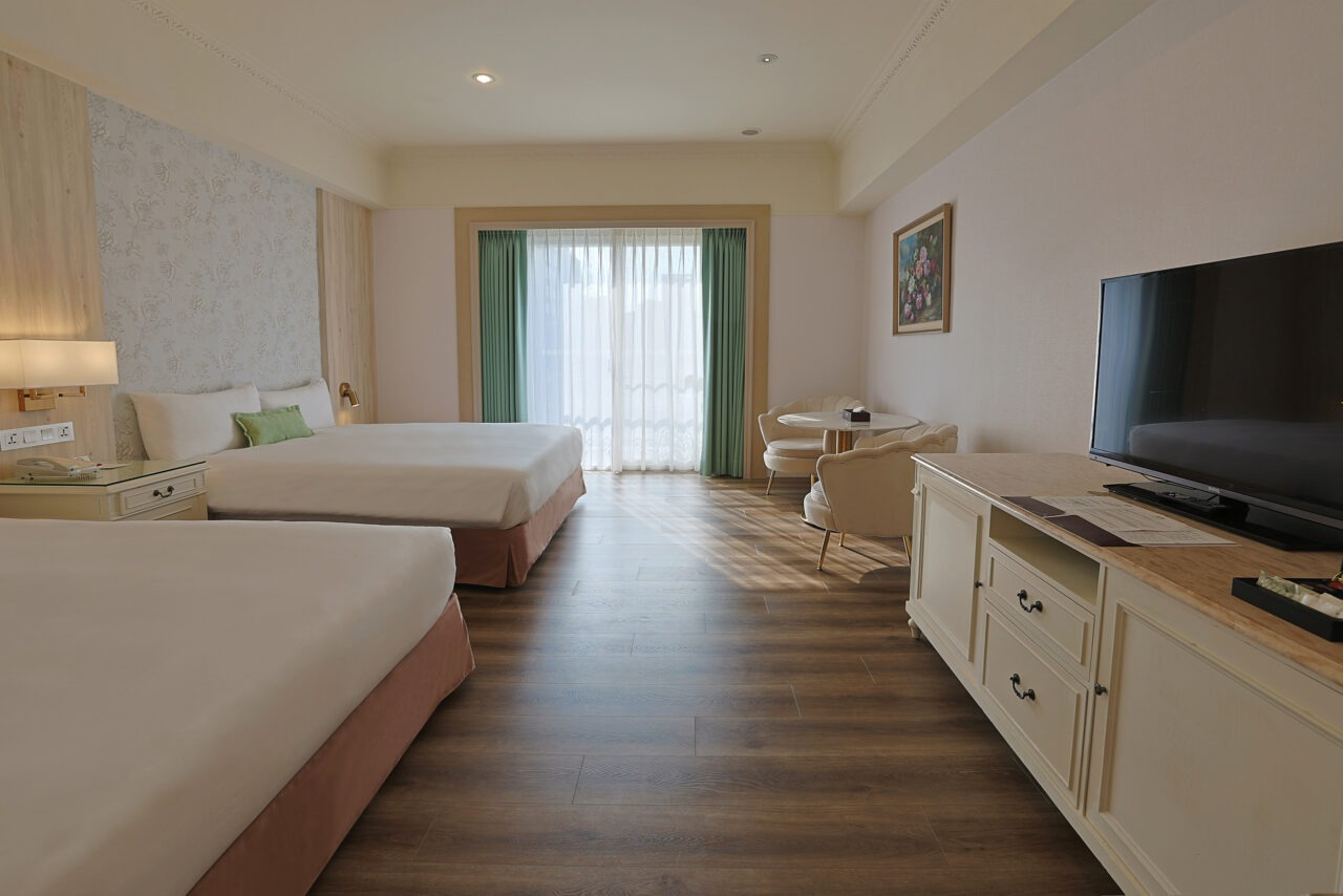 A spacious hotel room with two large beds, a TV console, and a bright balcony.