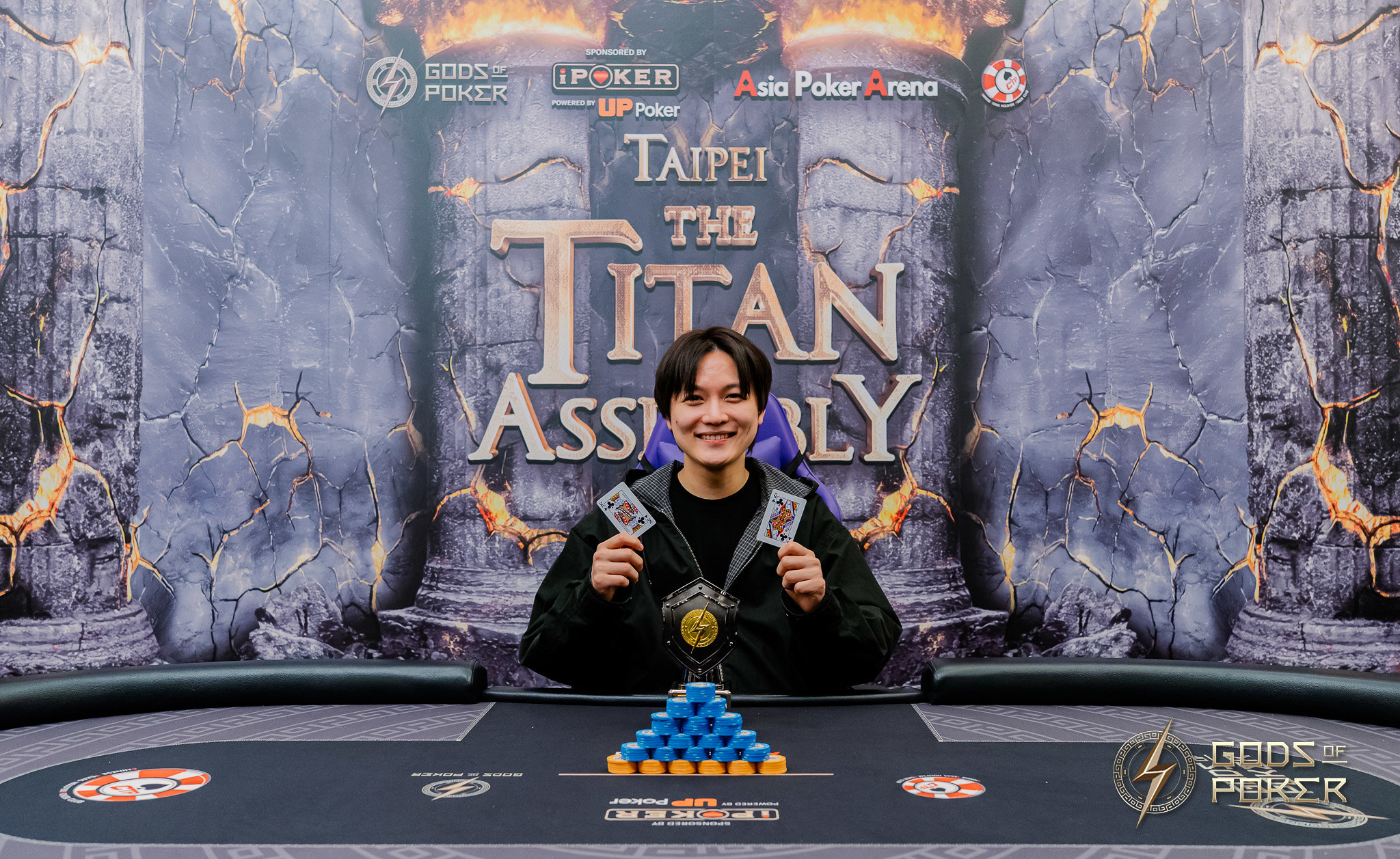 Smiling poker player holds up winning cards and a medal at 'The Titan Assembly'.