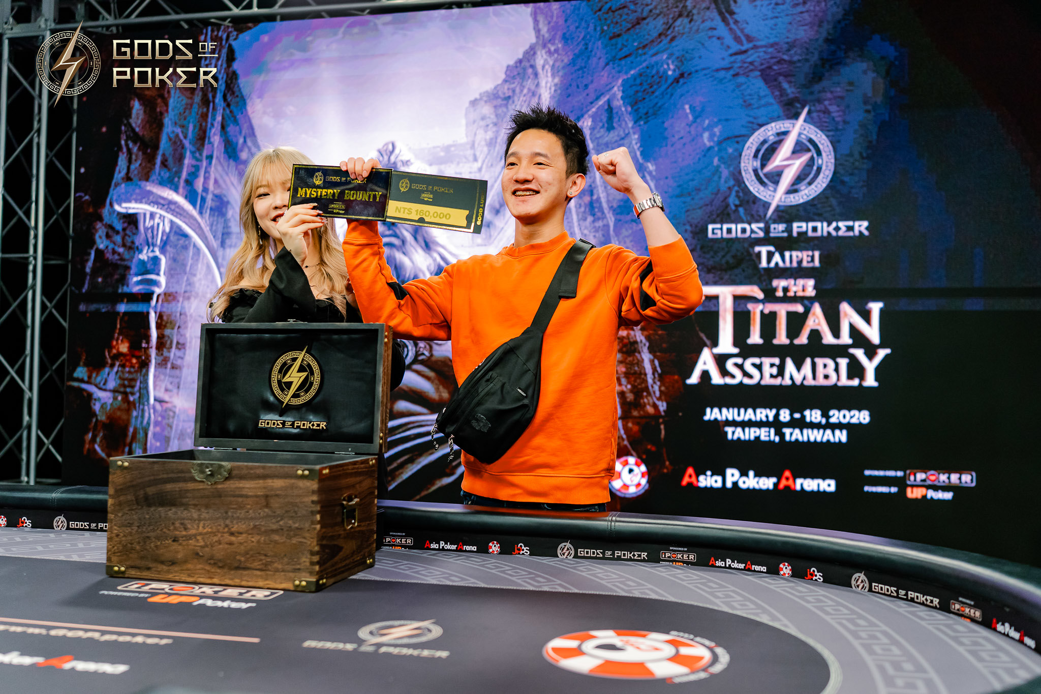 Man in orange shirt celebrates poker win with trophy, woman smiles beside a treasure box.