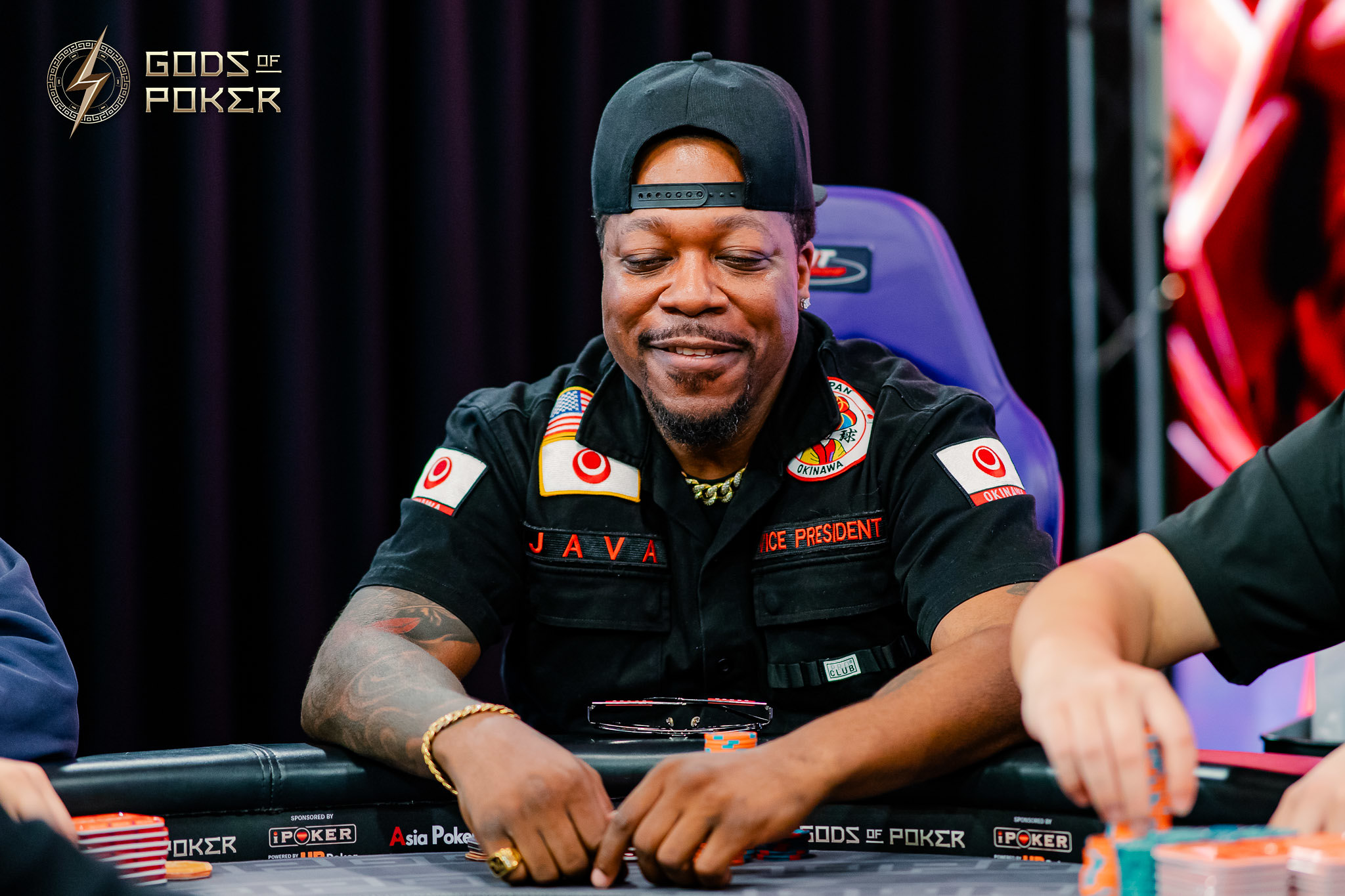 Smiling man in a black shirt and cap, playing poker at a table with chips.