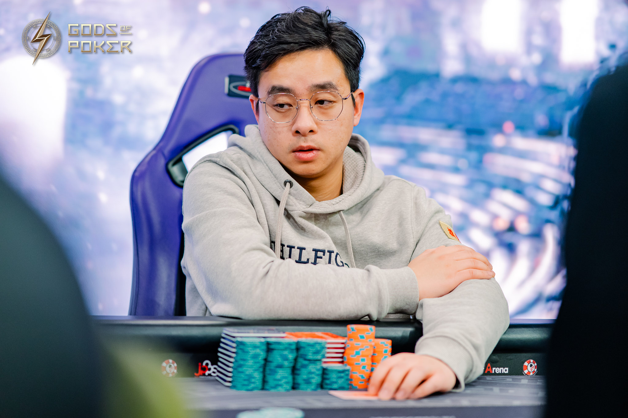 Young Asian man in glasses at poker table with chip stacks, looking thoughtful.