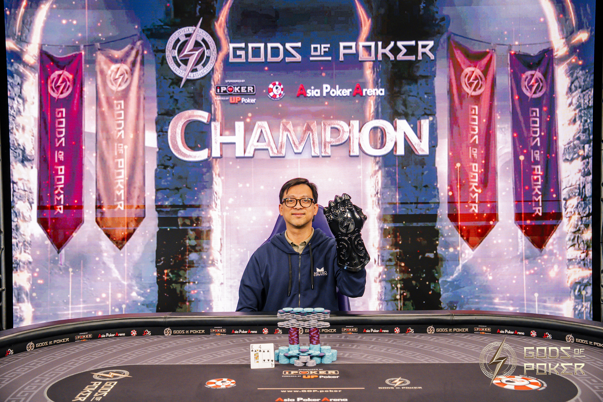 A man smiles, holding a trophy in front of a 'Gods of Poker Champion' screen.
