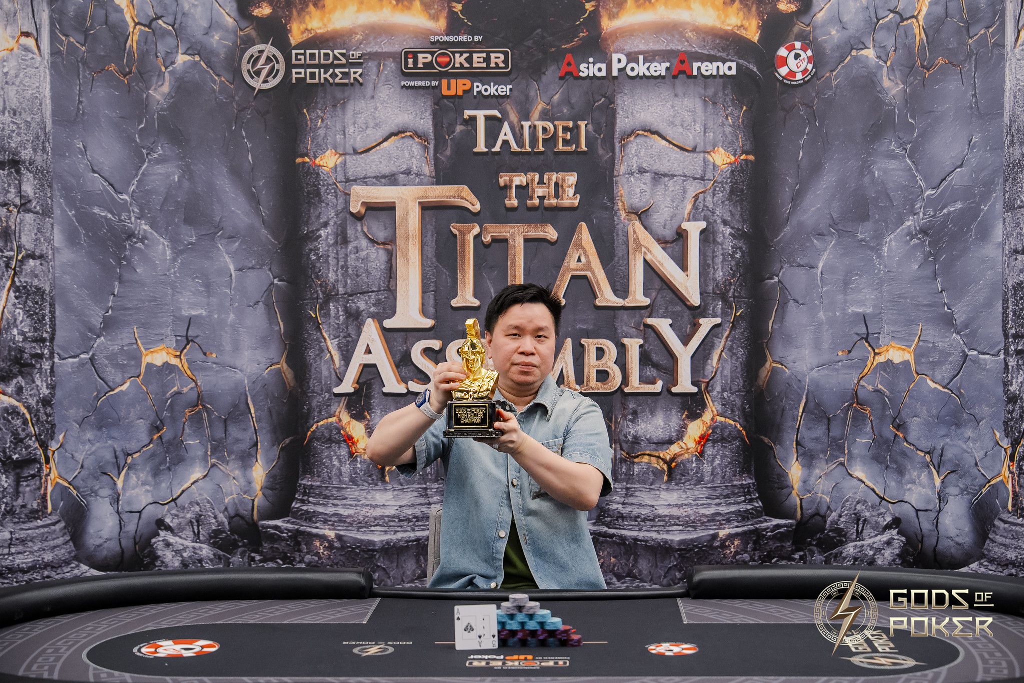 Man proudly holds a golden trophy at a poker table with a 'Titan Assembly' banner.