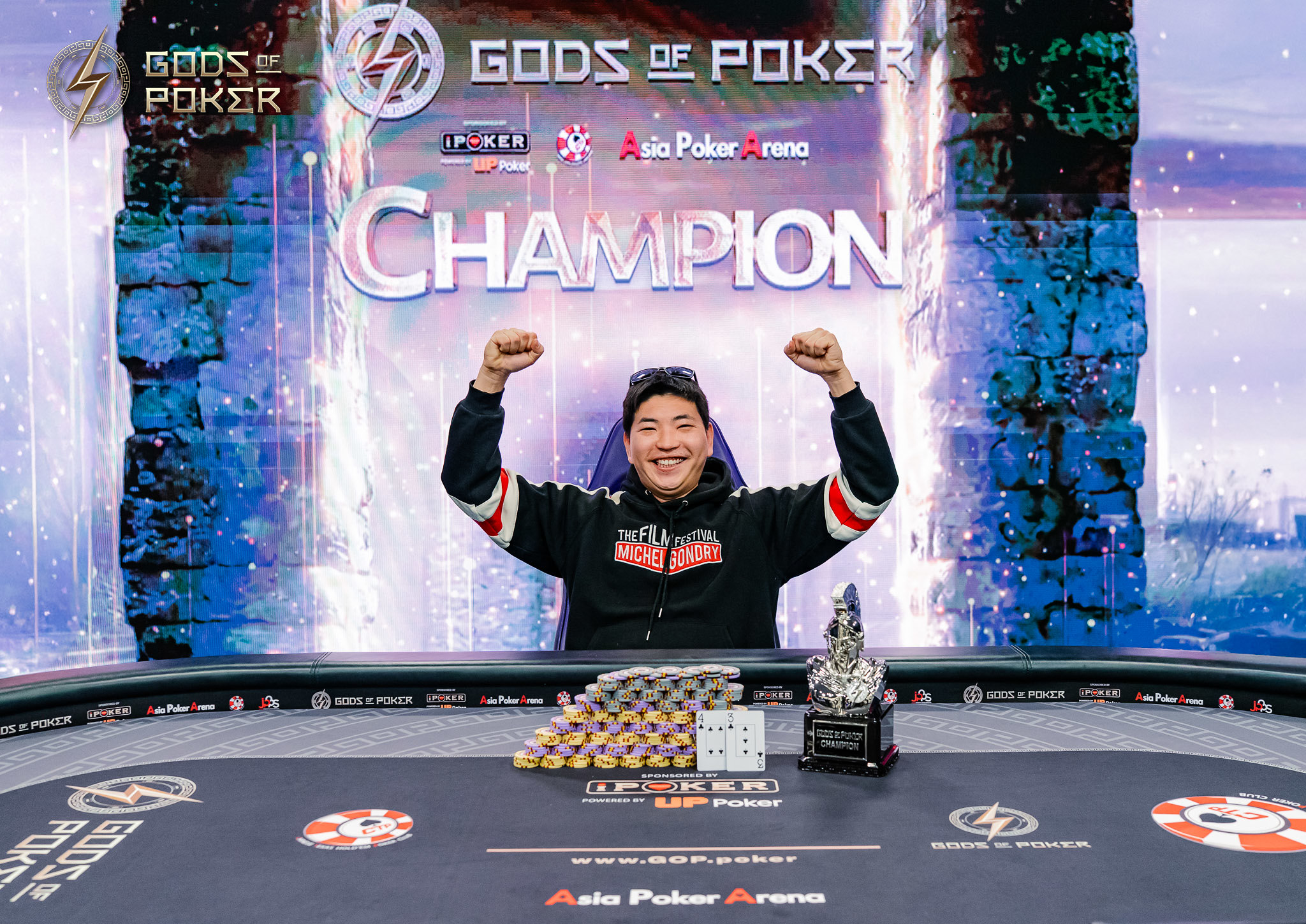 Smiling man raises arms in victory, celebrating a poker championship with a trophy and chips.